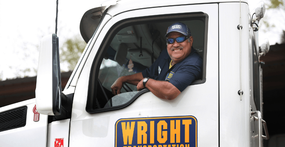 A truck driver is pictured leaning out of the window of a white 18-wheeler and smiling at the camera.