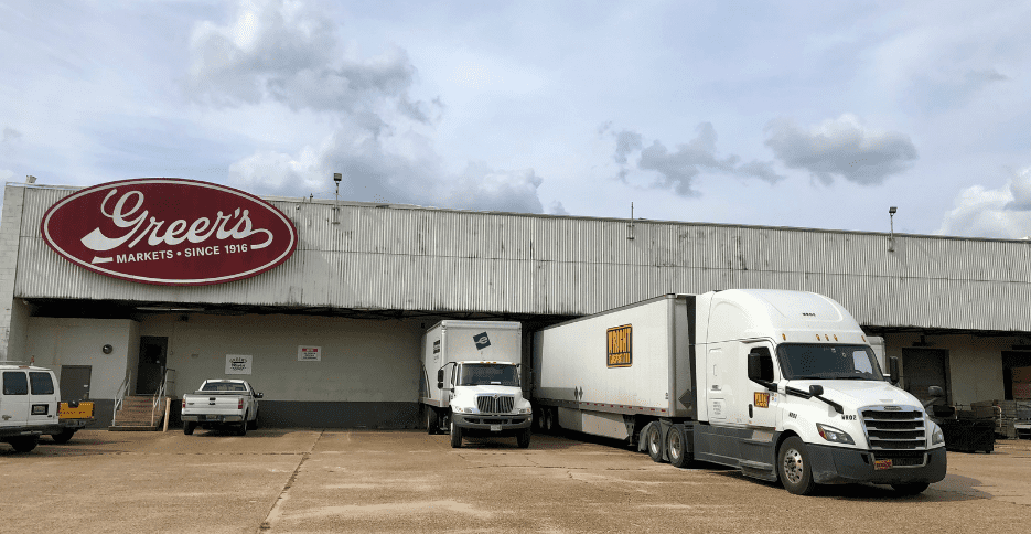 A white semi truck with the Wright Transportation logo is shown backed up to a loading dock at a Greer's Market warehouse.